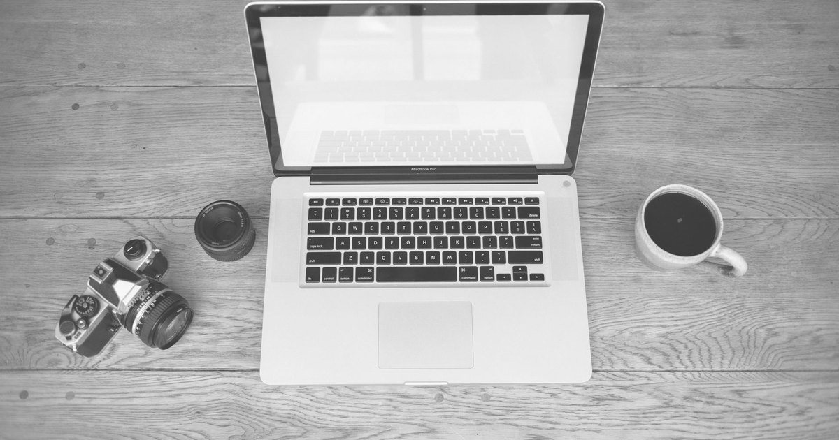 Monochrome image of a laptop, camera, lens, and coffee cup on a wooden desk