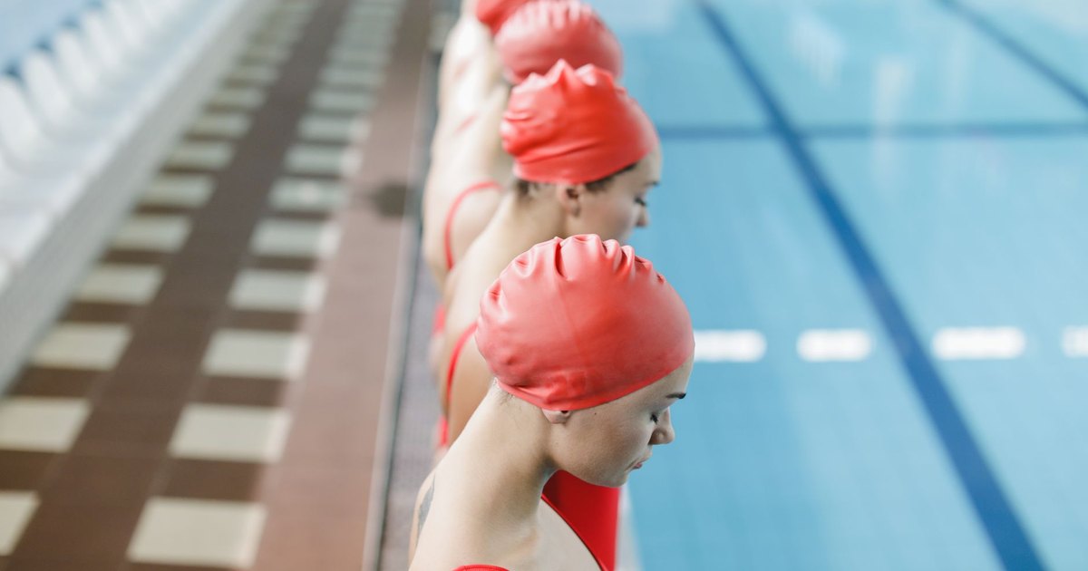 A team of female synchronized swimmers lined up by an indoor pool wearing red swimwear and caps.