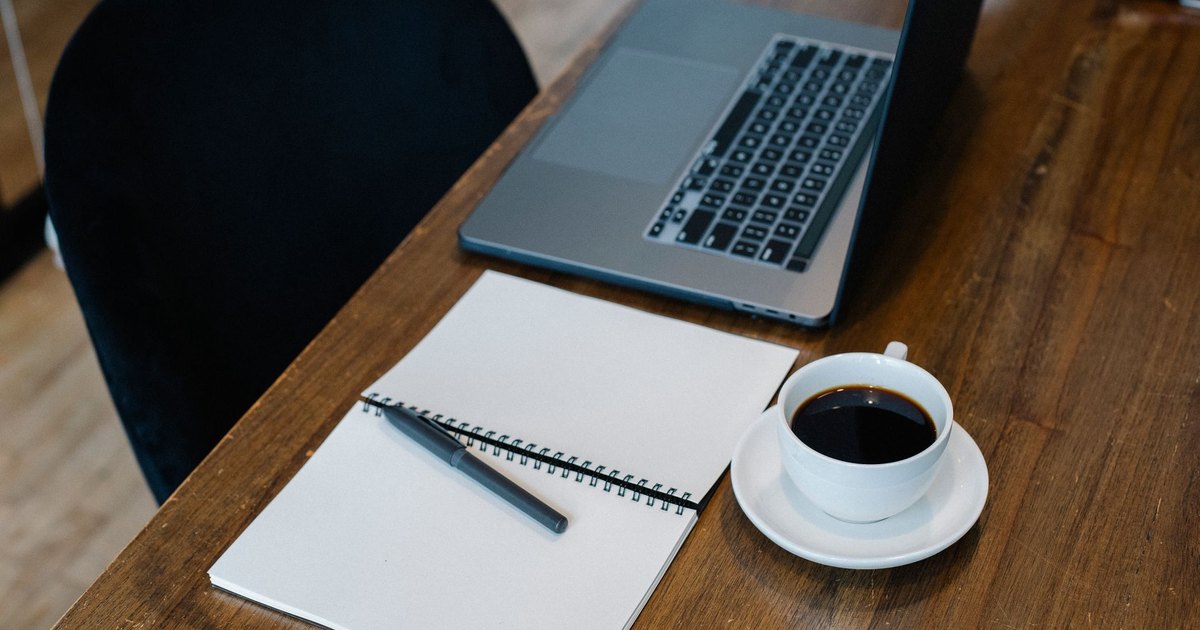 High angle of opened laptop and spiral notebook with blank pages placed on wooden table with cup of coffee and pen during workday