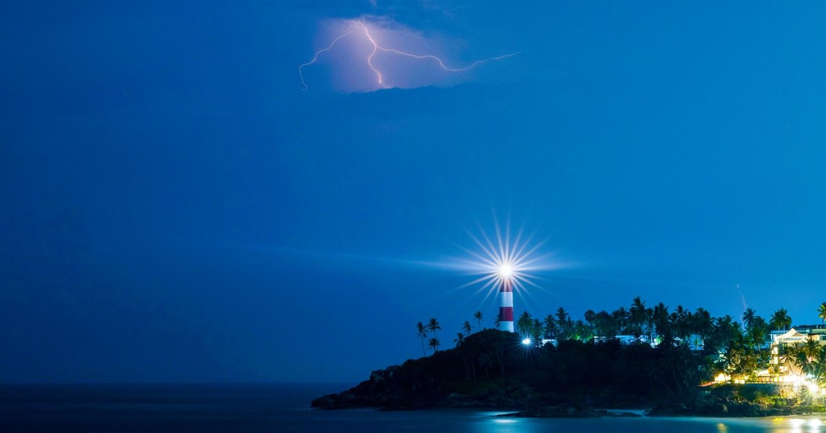Dramatic lighthouse illuminated at night with lightning over a coastal landscape.