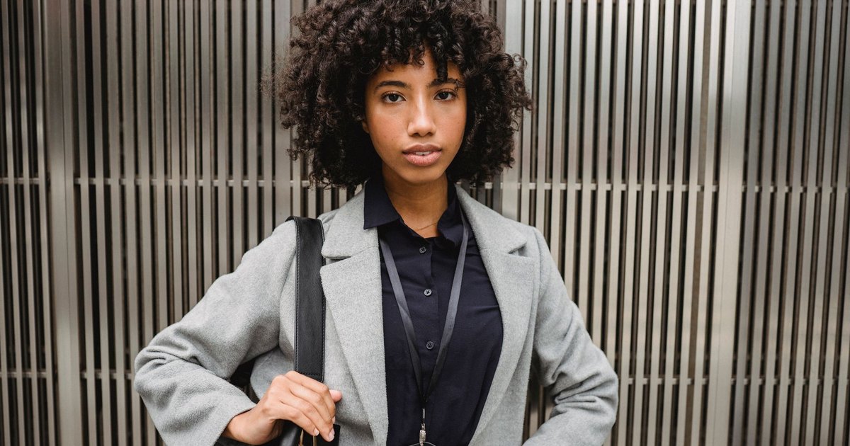 Elegant young woman with curly hair poses confidently in a gray coat against a modern metal background.
